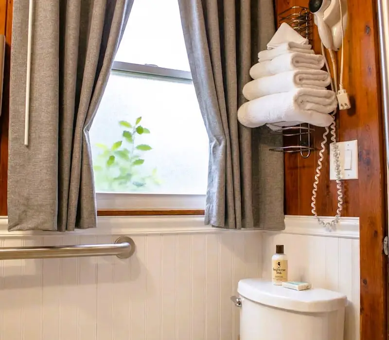 A bathroom with a toilet, a grab bar, a window with grey curtains, and a wall-mounted hairdryer and towel rack