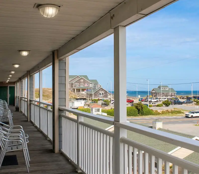 A covered outdoor walkway with white railings and chairs overlooks a street, parking lot, and buildings with the ocean in the background
