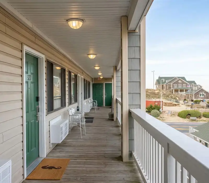 A long outdoor corridor with green doors and windows on the left, and a white railing on the right overlooking distant buildings