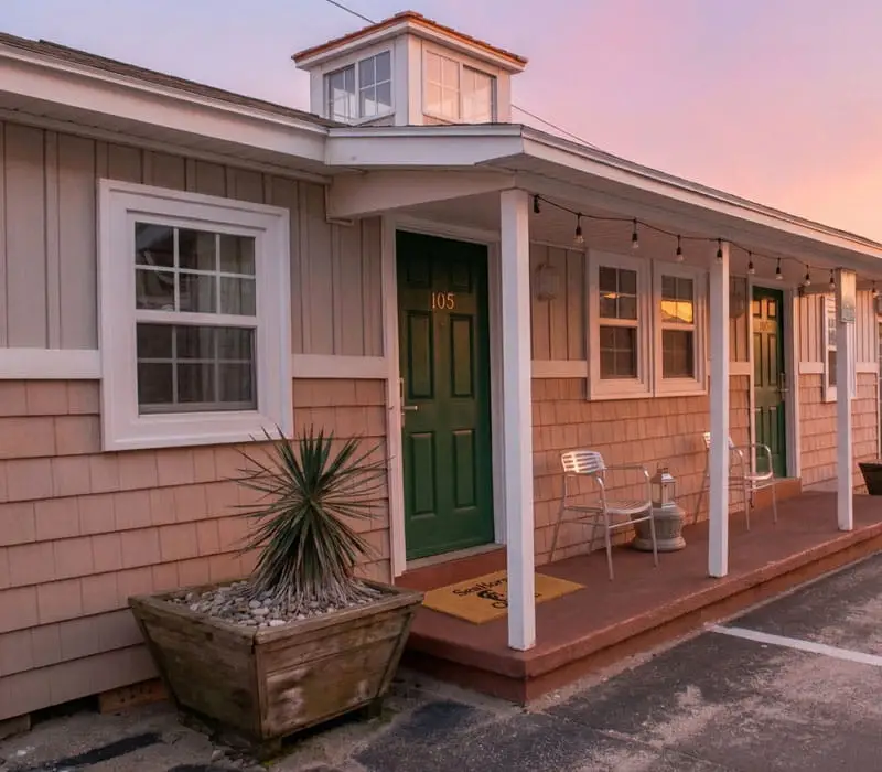 A single-storey building with light brown shingle siding, a dark green door numbered 105 and a small porch with two chairs