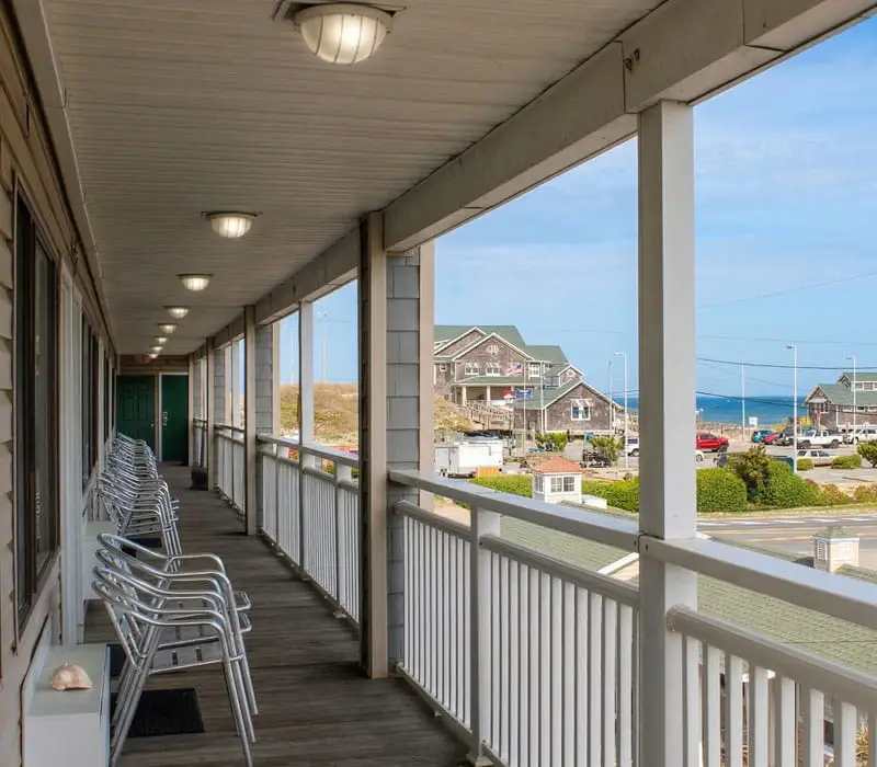 A long outdoor balcony with a row of chairs and a view of houses, a road and the ocean in the distance
