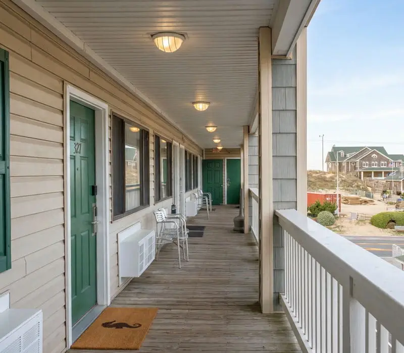 A long outdoor wooden walkway with green doors and windows on the left, and a white railing on the right overlooking a sandy area and buildings