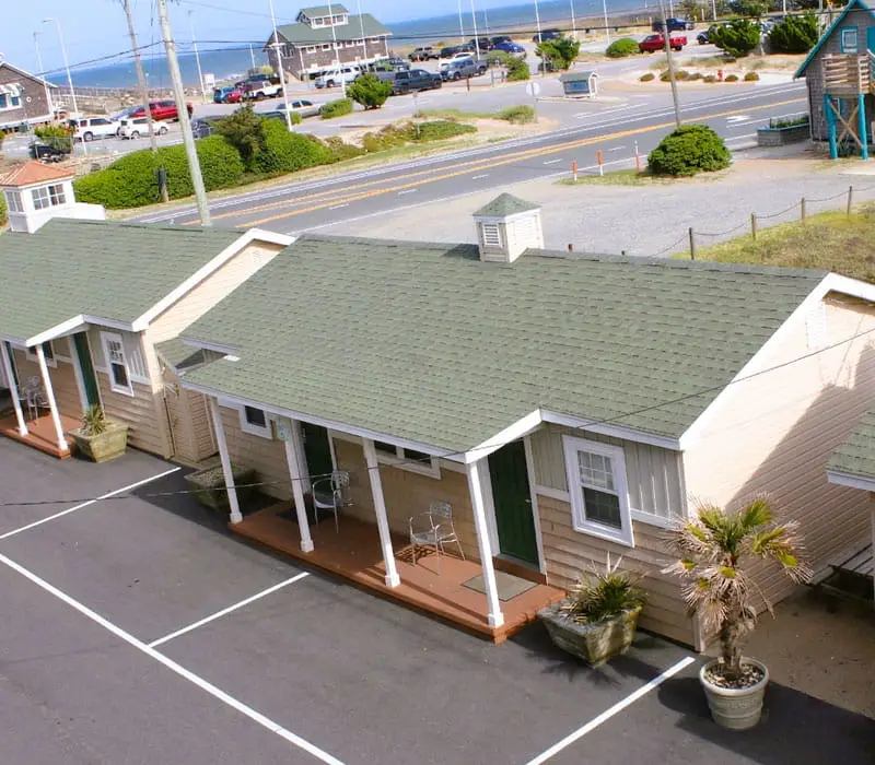 Two single-storey cabins with green roofs and front porches face a car park, with a road and the ocean in the background