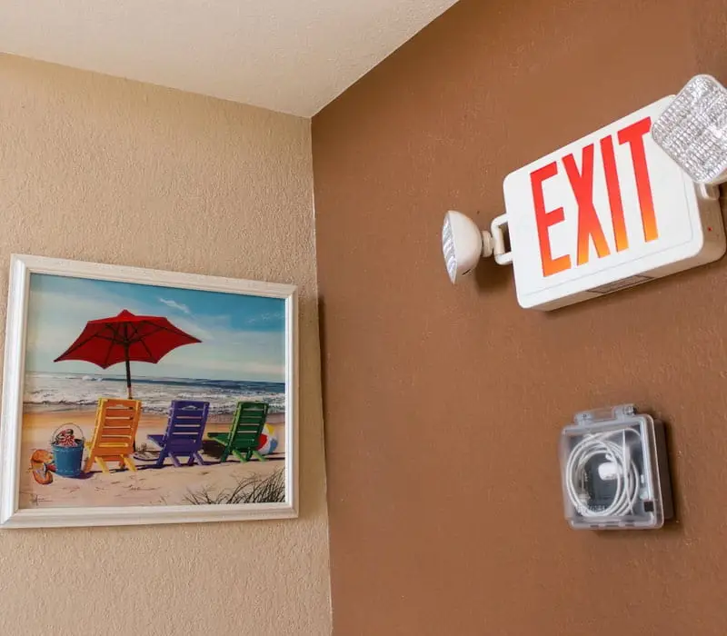 A corner of a hallway with a framed painting of a beach scene and an illuminated EXIT sign above a small clear box