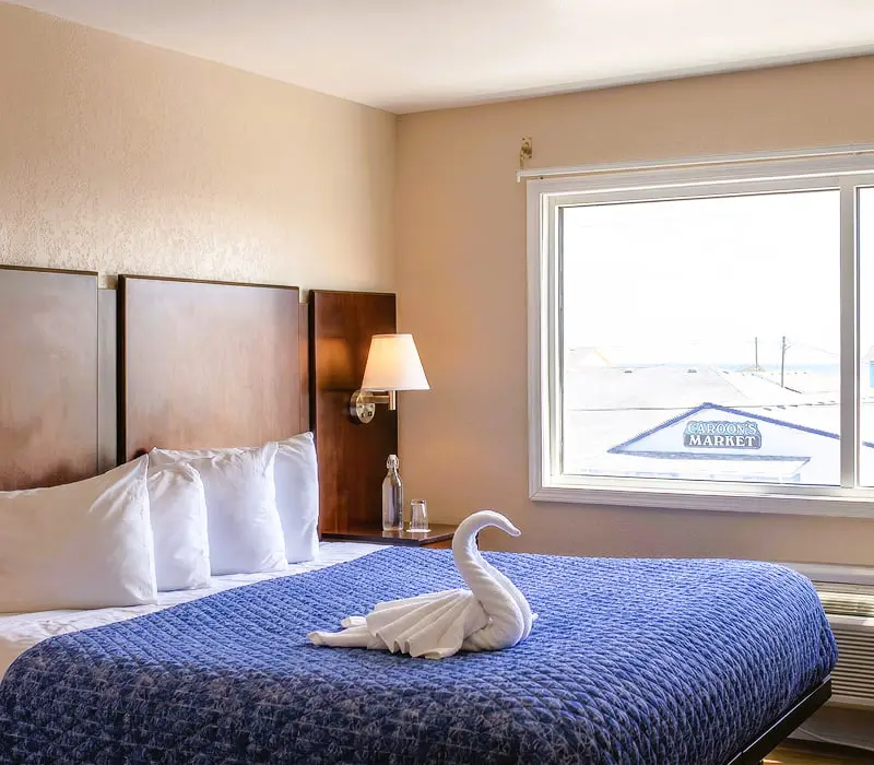 A king bed with a folded-towel swan beside a window overlooking neighbouring shop rooftops in a Premium Room at Seahorse Inn & Cottages