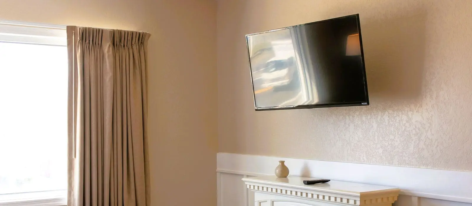 A wall-mounted television is above a white decorative fireplace with a small vase and a remote control