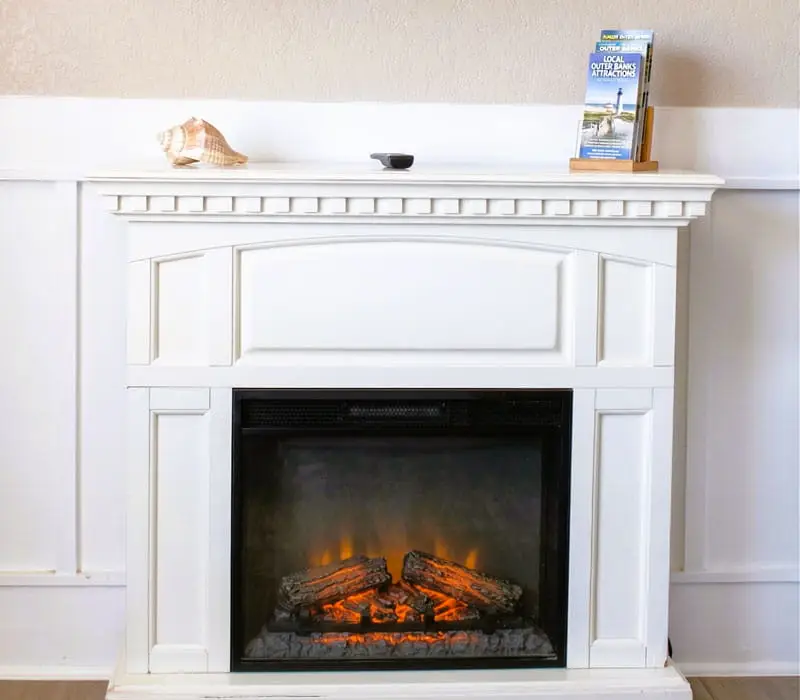 A white fireplace with an electric fire and a mantelpiece displaying a seashell and a brochure holder with "LOCAL OUTER BANKS ATTRACTIONS" visible