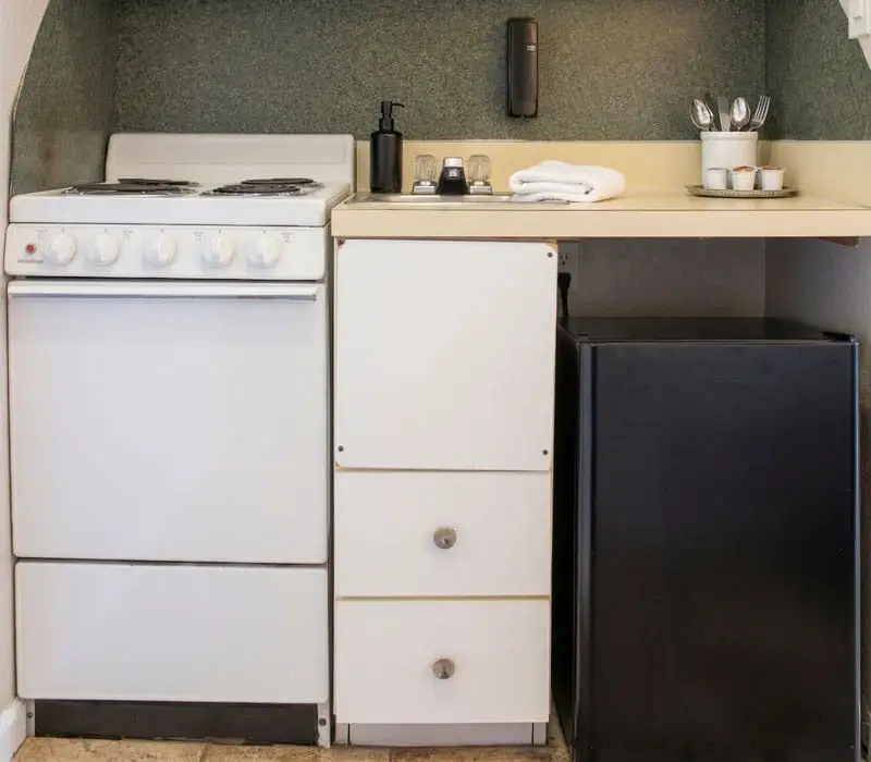 A kitchenette with a white electric stove, white drawers and a black mini-fridge under a counter with a sink