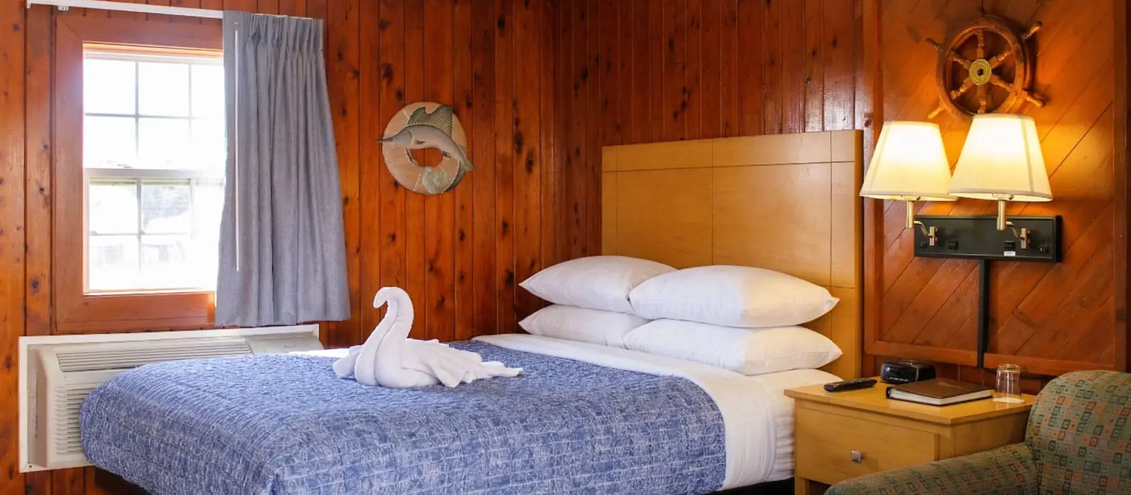 A guest room with wood-panelled walls, a bed with a blue patterned duvet, a window, and a bedside table with lamps