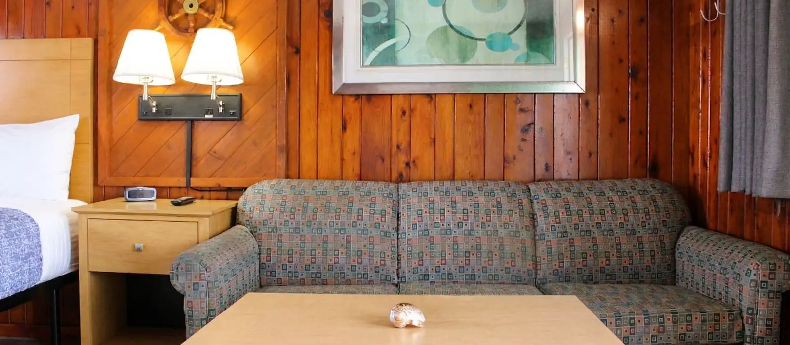 A guest room with a bed, a wooden bedside table, a patterned sofa, a wooden coffee table and wood-panelled walls
