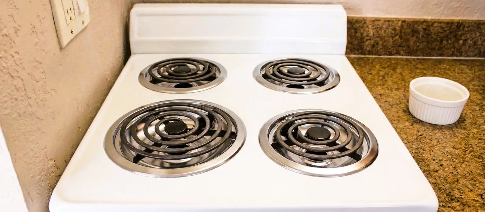 A white electric stove with four burners sits next to a speckled brown countertop with a small white bowl