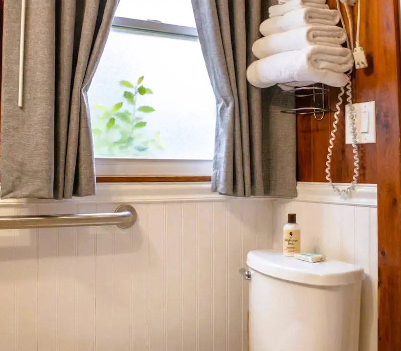 A bathroom with a toilet, a grab bar on the wall, a window with grey curtains, and a towel rack with folded white towels
