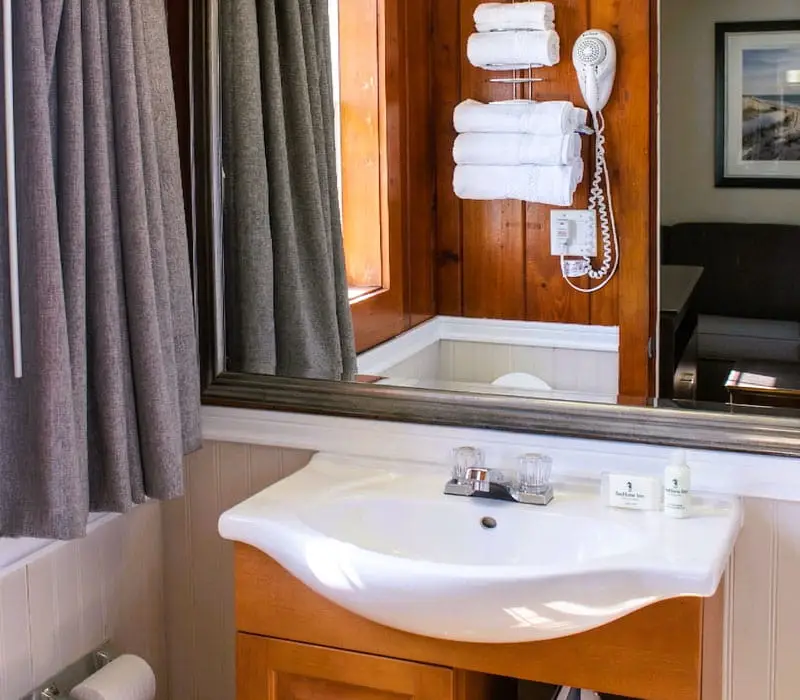 A bathroom vanity with a white sink, a mirror reflecting a window with grey curtains, and a hairdryer and towels mounted on a wood-panelled wall