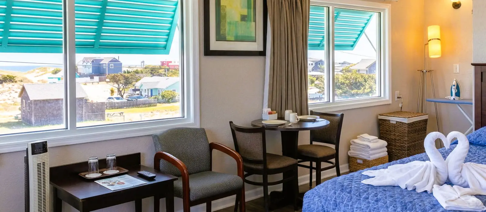 A hotel room with two large windows showing an ocean view, a small dining table with two chairs, and a bed with towel swans