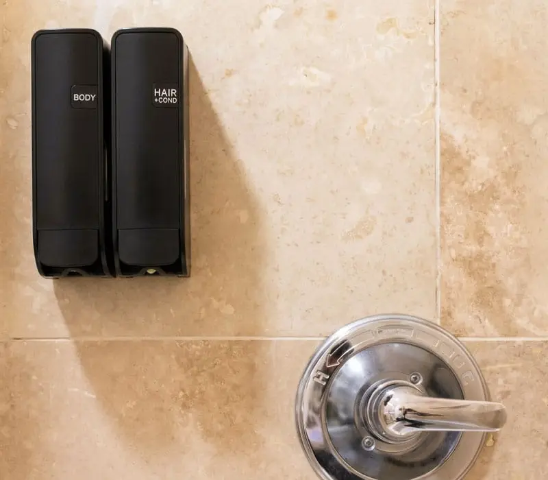Two black wall-mounted dispensers labelled "BODY" and "HAIR + COND" are above a chrome shower control on a tiled wall