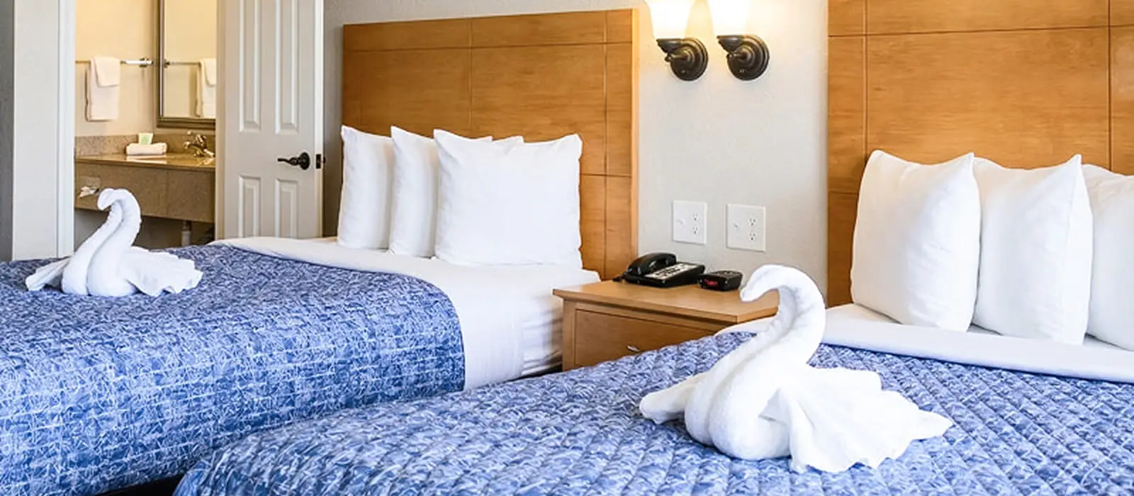 A guest room with two beds with blue patterned bedspreads, white pillows and towel swans, and a partial view of a bathroom